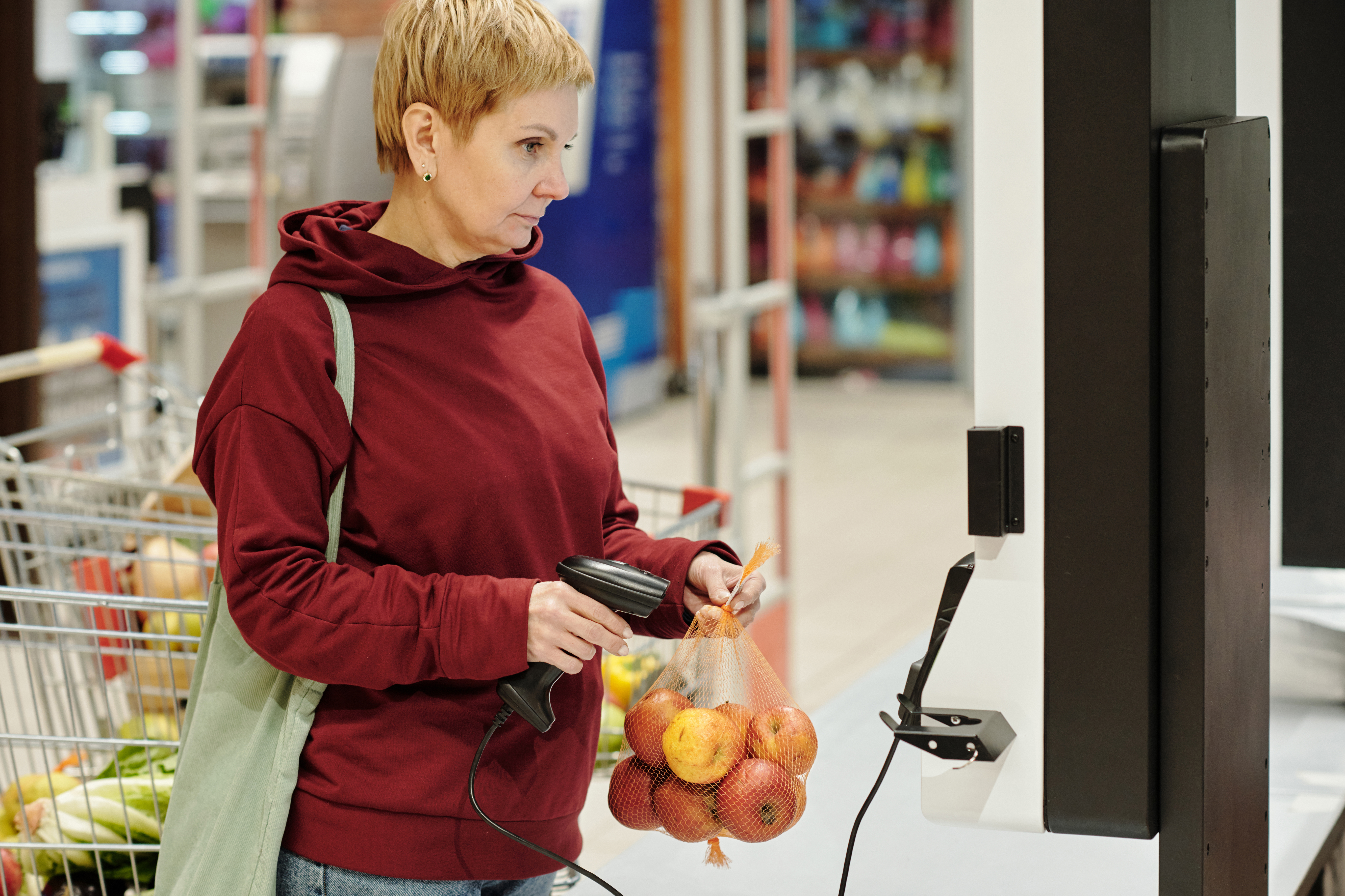 Blond mature female consumer in casualwear looking at screen with price of product while scanning pack of fresh ripe apples in supermarket