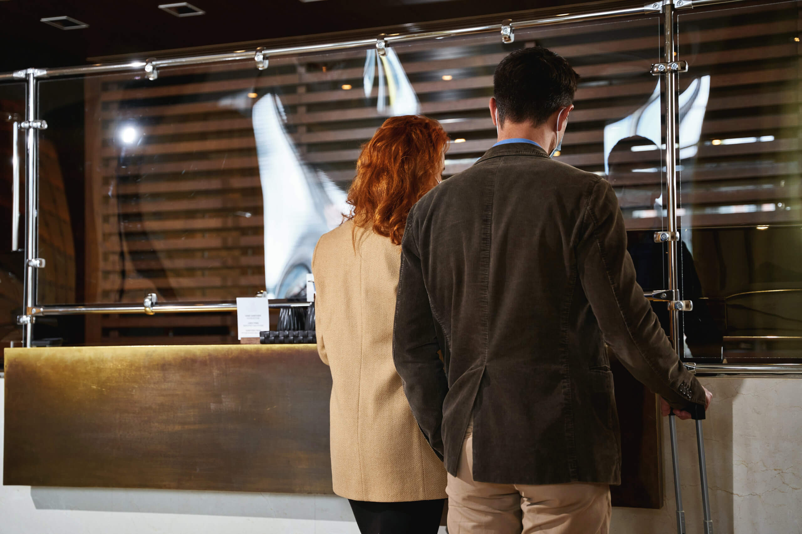 Rear view of an adult couple standing in front of a glass shield on the countertop during the check-in. Template banner